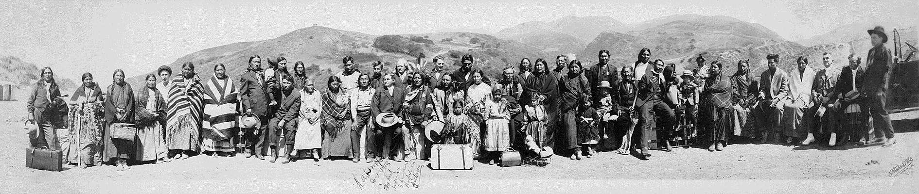 This black and white image depicts a gathering of roughly forty Native Americans gathered for a group photo in front of low mountains.