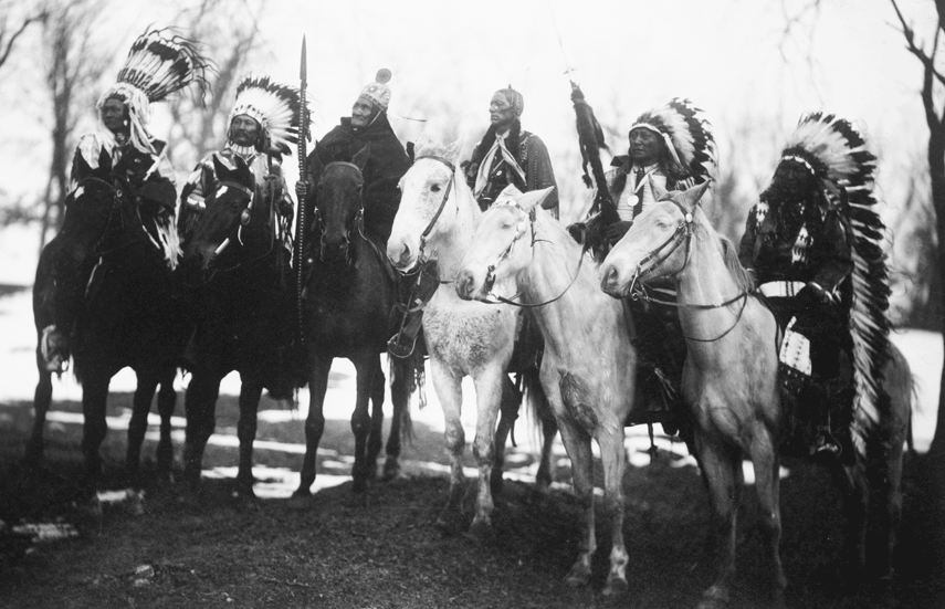 Six Native American tribal leaders sit on horseback. This image is in black and white.